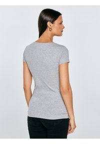 Kobieta o prostych ciemnych włosach stoi bokiem, zwrócona w prawo, ubrana w dopasowany szary t-shirt z krótkim rękawem i czarne spodnie na jednolitym tle.