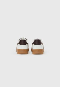 Une paire de baskets blanches avec des empiècements en cuir marron sur le talon et des semelles en caoutchouc caramel, vue de l'arrière sur un fond blanc.
