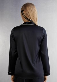 Långärmad marinblå satinblus med en slät textur, ljus kantning och en klassisk kragsdesign, sedd bakifrån.