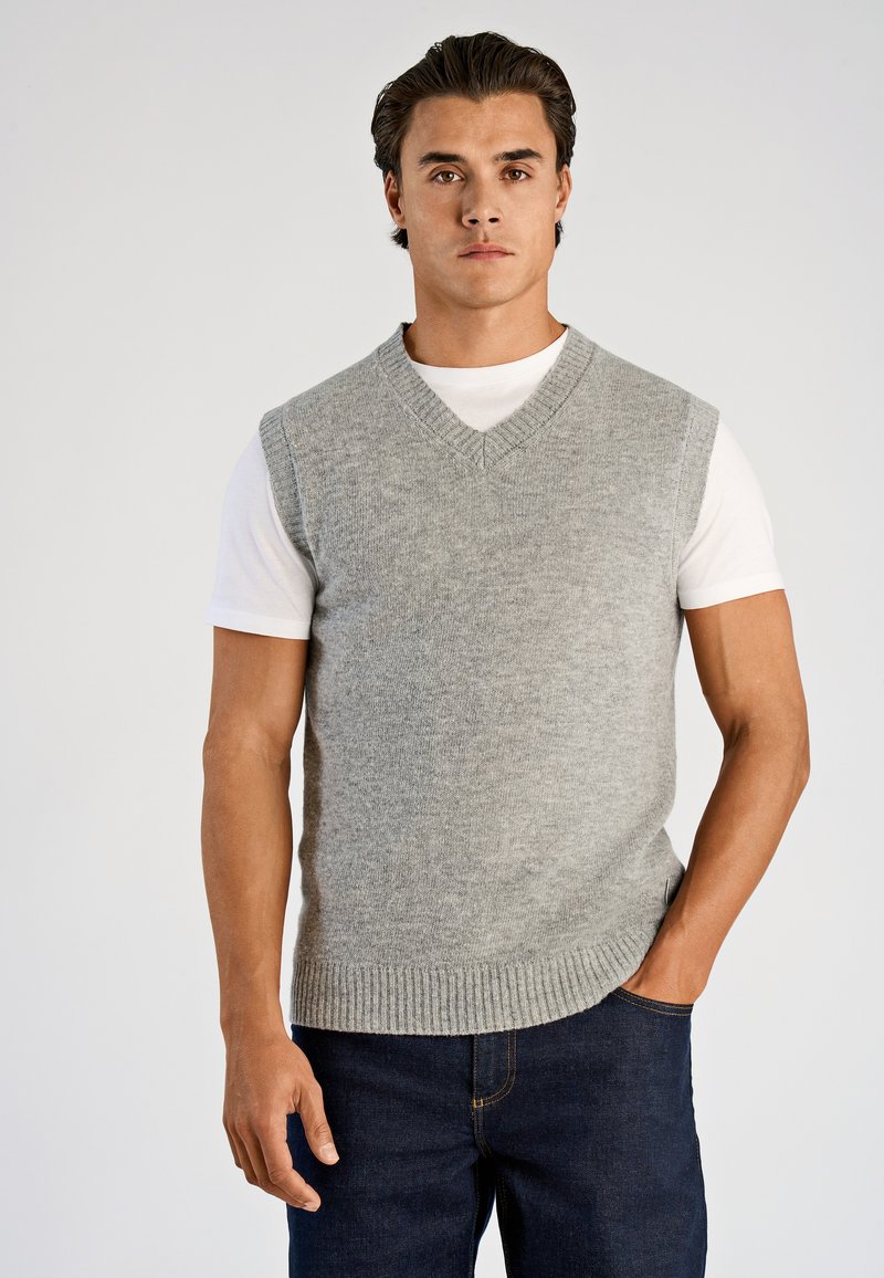 Grå stickad ärmlös tröja med V-ringning. Har ribbad fåll och kombineras med en vit t-shirt och mörka jeans.