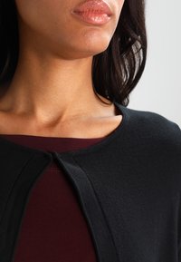 Cardigan negro con un cuello redondo y un pequeño cierre frontal, superpuesto a una blusa color granate. La tela parece suave y tiene una textura agradable.