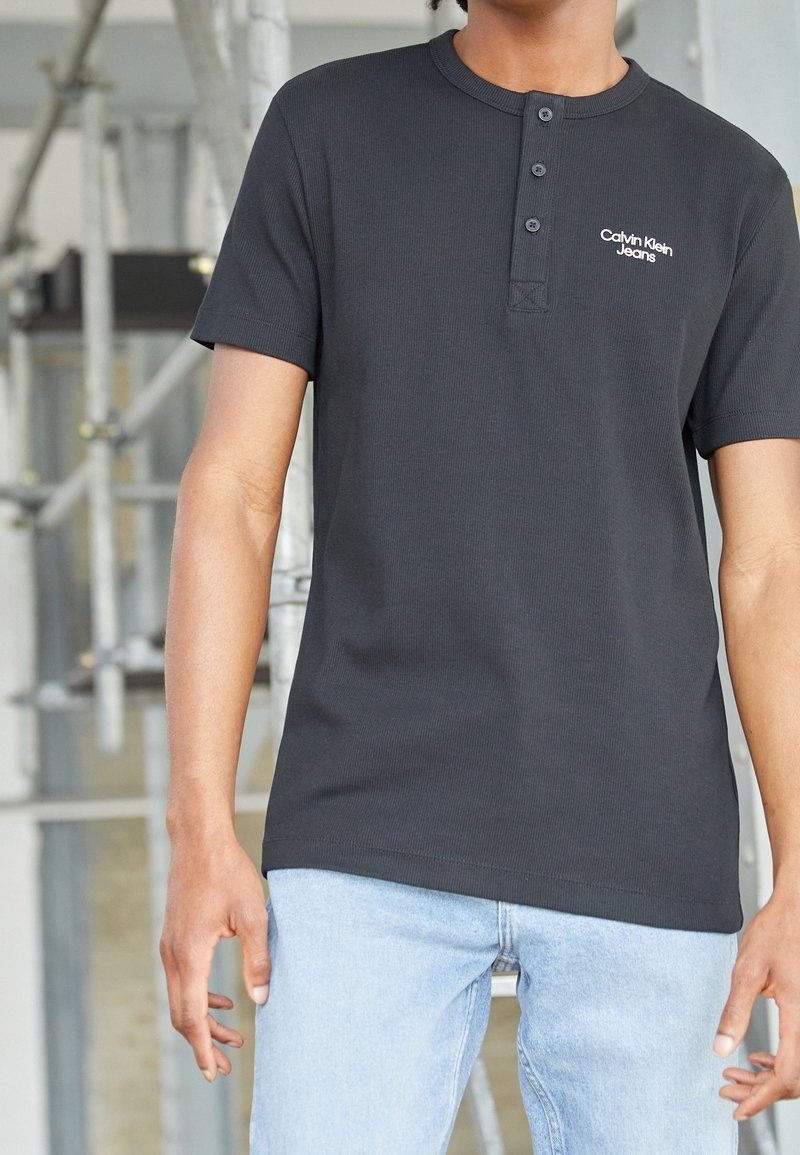 Černé krátkosrsté henley tričko z žebrované tkaniny s klopou na tři knoflíky a vyšitým logem "Calvin Klein Jeans" na levé hrudi.