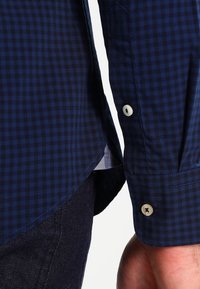 Primer plano de una mano al lado del puño de una camisa a cuadros azul oscuro con botones blancos, combinada con jeans de mezclilla oscuros.