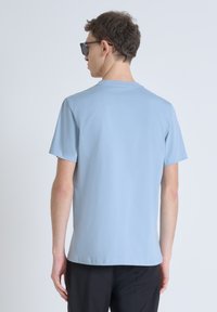 Camiseta de algodón azul claro con mangas cortas, cuello redondo, corte recto y textura suave. Llevada con pantalones oscuros; vista trasera.