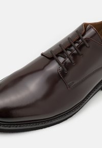 Brune lærdressko med en glansfull finish, som har seks hull og mørke snører, rund tå og strukturet såle.