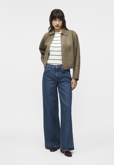 Žena s tmavými vlasmi v hnedej semišovej bunde, bielej pruhovanej košeli, džínsach s širokými nohavicami a tmavými topánkami stojaca pred bielym pozadím.