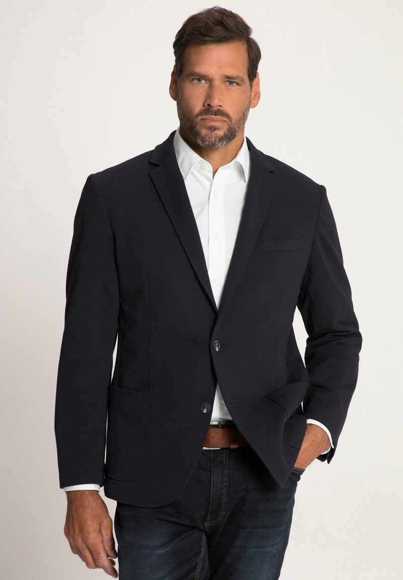 Marineblazer mit taillierter Passform, ausgestattet mit zwei Fronttaschen, einer Brusttasche und zwei Knöpfen. Getragen über einem weißen Hemd und dunklen Jeans.