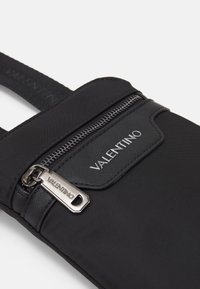 Sac en nylon noir avec une poche zippée à l'avant, logo de la marque en relief et bretelles réglables. Les détails en métal argenté améliorent la fonctionnalité.
