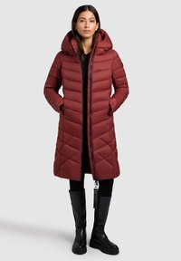 Červená puffa bunda s kapucňou, s prešívaným dizajnom, zip uzáverom a bočnými vreckami, spárovaná s čiernymi vysokými čižmami.