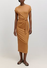 Robe beige jusqu'aux genoux avec des manches courtes et un détail à nouer à la taille. Tissu lisse avec une silhouette ajustée. Associée à des sandales noires à lanières.