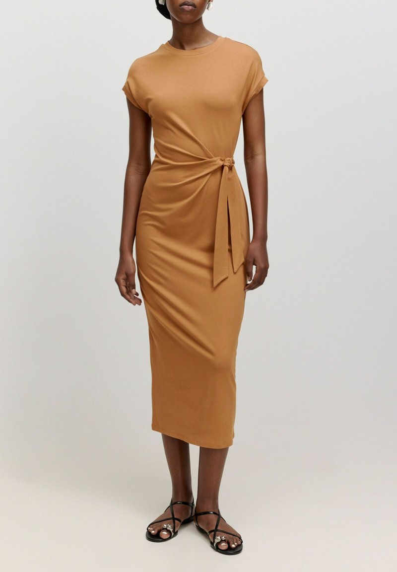 Robe beige jusqu'aux genoux avec des manches courtes et un détail à nouer à la taille. Tissu lisse avec une silhouette ajustée. Associée à des sandales noires à lanières.