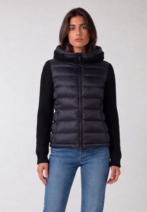 Vrouw draagt een zwarte doorgestikte bodywarmer met gebreide lange mouwen en kraag, gecombineerd met blauwe jeans, staand tegen een effen achtergrond.