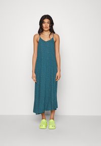 Robe maxi teal avec un décolleté en V, des fines bretelles réglables et un petit motif de points blancs ; associée à des sabots verts vif.