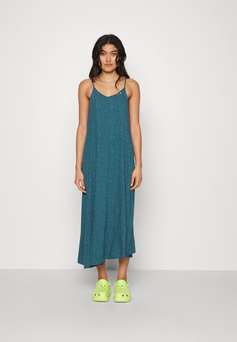 Robe maxi teal avec un décolleté en V, des fines bretelles réglables et un petit motif de points blancs ; associée à des sabots verts vif.