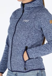 Giacca in pile con zip di colore blu navy, tessuto testurizzato, colletto alto, tasche laterali e bordi di un blu scuro a contrasto. Toppa del logo sulla spalla.