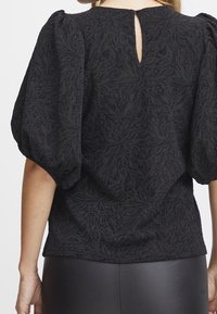 Blusa nera in tessuto testurizzato con maniche a sbuffo e apertura a chiave sul retro. Motivo floreale dettagliato su tutto il tessuto.