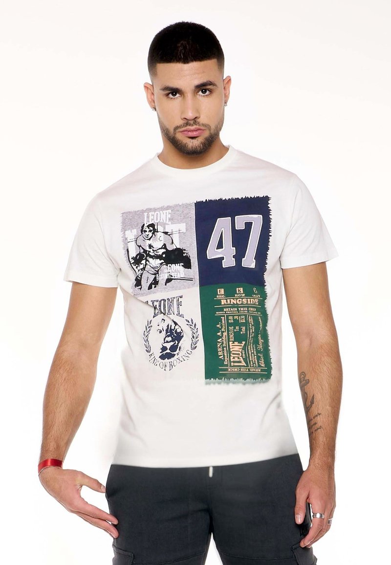 Homme portant un t-shirt blanc avec un graphisme sur le thème de la boxe, numéro 47 et texte, associé à un pantalon foncé sur fond blanc uni.