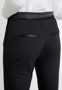Schwarze, maßgeschneiderte Hosen mit glatter Textur, satinierter Taille und zwei dekorativen Gesäßtaschen mit satiniertem Rand.