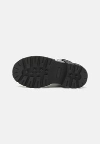 Semelle de botte en caoutchouc noire avec de forts motifs de crampons, des zones surélevées et une section lisse. Présente une découpe rectangulaire pour le marquage de la marque.