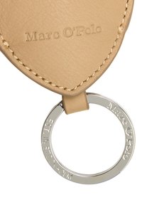 Beżowy skórzany brelok z wytłoczonym logo "Marc O'Polo" i srebrnym metalowym pierścieniem z wygrawerowanym napisem. Widoczne detale teksturowanego szwu.