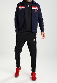 Marinblå träningsoverall med röda och vita ränder, zip-jacka, svart T-shirt, svarta byxor med vita sidostar och röda träningsskor.