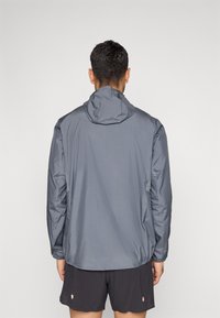 Γκρι αδιάβροχο ελαφρύ μπουφάν με κουκούλα, ελαστικά μανίκια και λείο ύφασμα ανθεκτικό στο νερό. Μινιμαλιστικός σχεδιασμός, χωρίς ορατά λογότυπα ή σχέδια.