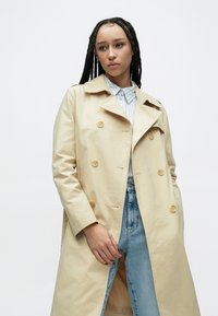 Béžový trenčkot s dvojradovým zapínaním, žltými gombíkmi, širokým golierom a bočnými vreckami. Nosený cez svetlomodrú košeľu na gombíky a rifle.