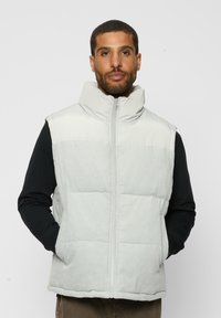 Chaleco acolchado en corduroy gris claro con cuello alto, cierre frontal y bolsillos laterales; superpuesto a una camisa de manga larga negra.