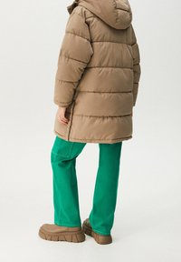 Manteau matelassé marron avec quilting horizontal, pantalon cargo vert, et chaussures marron épaisses. Capuche et poignets visibles, design minimaliste.