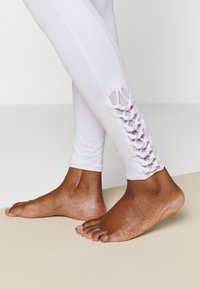 Leggings de um tom roxo claro com um painel lateral de malha respirável apresentando um cordão em cruz. Textura suave e design justinho. Descalça em um tapete de ioga.