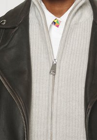 Braune Lederjacke über einem hellgrauen strukturierten Zip-Hoodie mit einem silbernen Reißverschluss. Der Kragen des weißen Shirts hat ein mehrfarbiges geometrisches Detail.