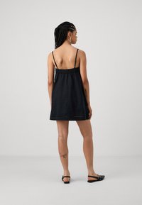 Schwarzes Leinen-Mini-Kleid mit elastischem Taillenbund und dünnen Trägern, gerade geschnitten und mit subtilen Saumdetails. Tragen mit schwarzen flachen Schuhen.