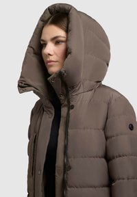 Ung kvinde iført en brun polstret vinterjakke med en stor hætte, lynet og knappet op, mod en ensfarvet lys baggrund.