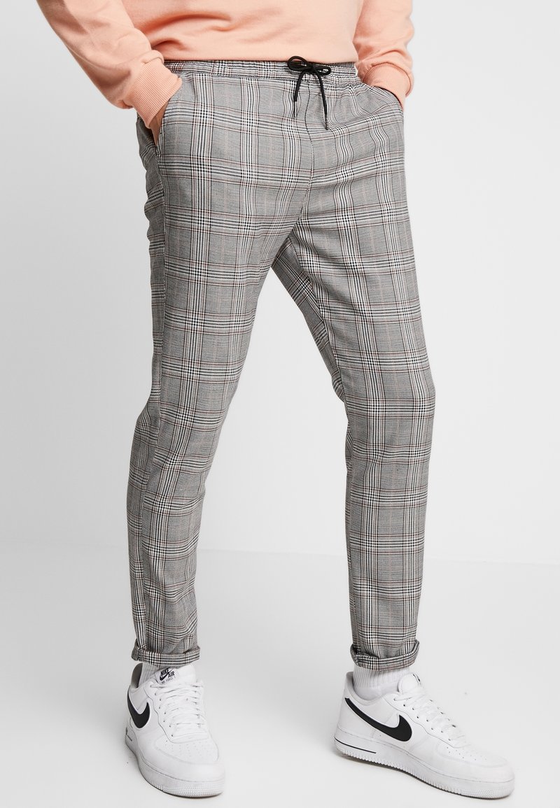Personne portant un pantalon à carreaux gris avec cordon de serrage, un sweat-shirt pêche, des baskets blanches Nike, les mains dans les poches, debout sur un fond blanc.