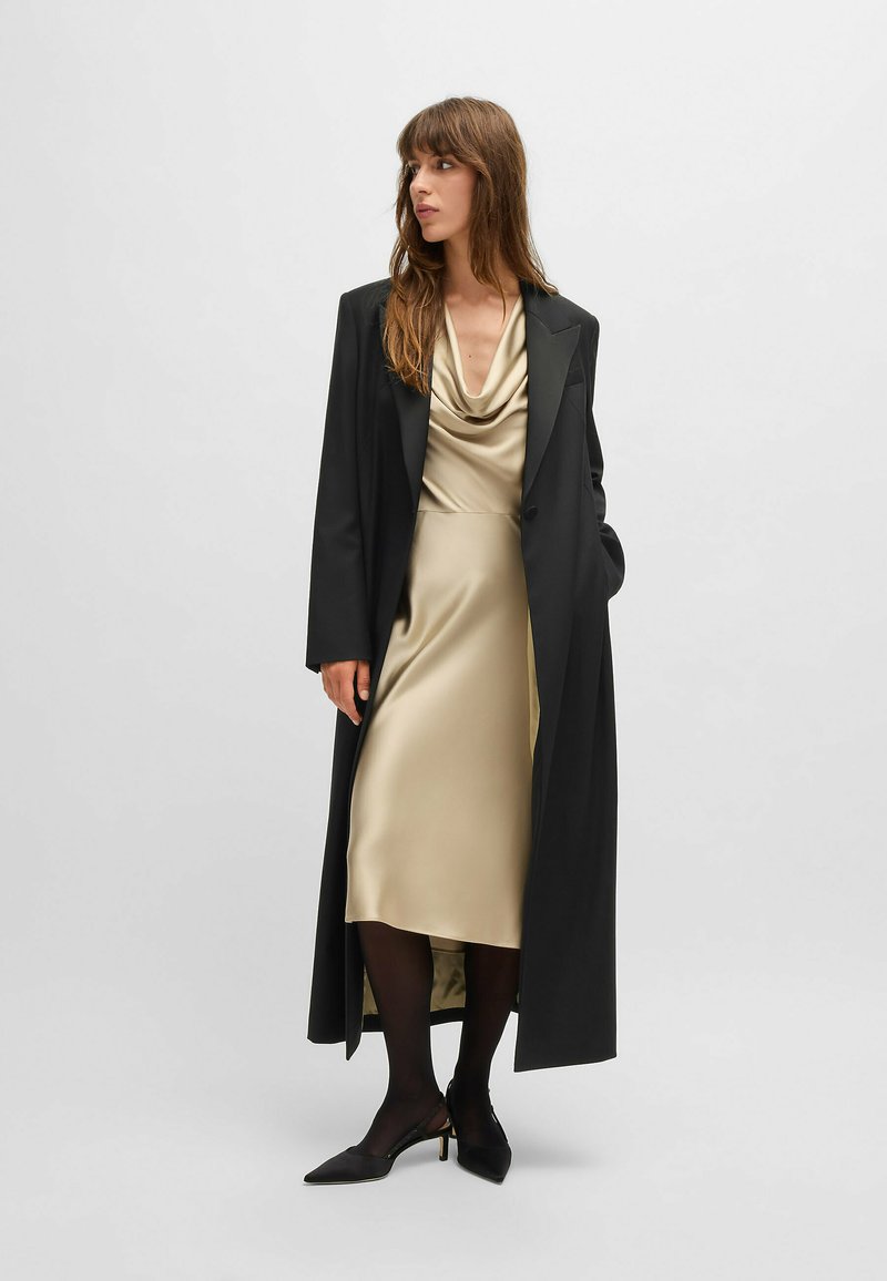 Manteau long noir porté sur une robe satin à col cheminée couleur or clair. La tenue est accompagnée de collants noirs et de talons aiguilles noirs, créant une silhouette épurée.