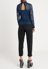 Blusa in pizzo navy con colletto alto e design con schiena scoperta, abbinata a pantaloni neri a sigaretta e sandali con tacco a fantasia a quadri.