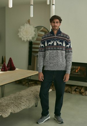 Strickpullover mit einem marineblauen und grauen Design, das Wintermotive zeigt, einem Reißverschlusskragen und gerippten Bündchen, kombiniert mit dunklen Jeans und grauen Sneakers.