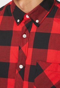 Camisa de cuadros roja y negra con botones, cuello abierto, botones blancos y un bolsillo en el pecho con tela texturizada.