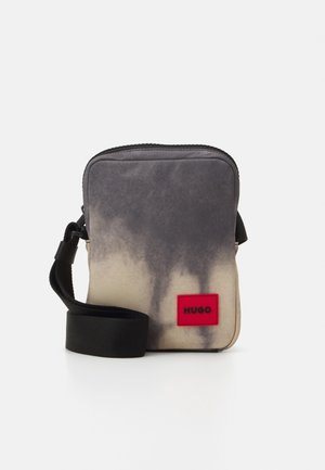 Lille rektangulær taske i en gradering fra grå til beige, med en sort lynlås, justerbar sort rem og en rød logo-patch.