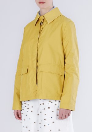 Kobieta ubrana w musztardową żółtą kurtkę z dużym kołnierzem i kieszeniami z patkami, w połączeniu z białą spódnicą ozdobioną małymi ćwiekami.