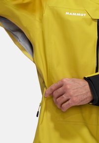 Giacca impermeabile gialla con tasca con zip. Presenta un design elegante e un tessuto texture. Accento nero nell'area del polso con fodera grigia.