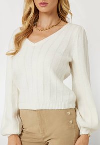 Suéter blanco de punto con escote en V, textura acanalada y mangas abullonadas. Combinado con pantalones de color beige que cuentan con detalles de botones dorados.