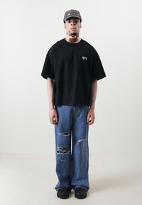 Crna oversized majica s logom, izlizane plave traperice s mrljama od boje i crne chunky cipele. Ležerni streetwear stil.