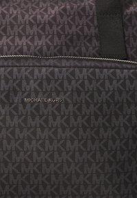 Sac en tissu noir avec un motif répétitif de logo "MK", une marque métallisée "Michael Kors" et des anses en nylon noir. Texture lisse.