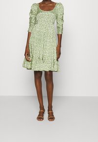 Green floral dress with puff sleeves and a square neckline, featuring a tiered hem and lightweight fabric. Paired with brown sandals.
