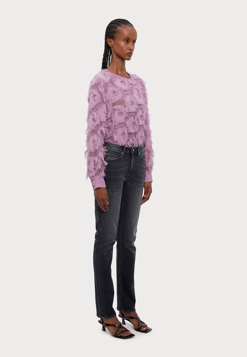 Suéter de color lila con flecos y mangas largas, combinado con pantalones skinny gris oscuro y sandalias de tacón negro con tiras. Elementos de diseño simples visibles.