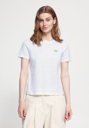 Kobieta o włosach do ramion, ubrana w biały krótki rękaw t-shirt z małym zielonym logo rakiet tenisowych i beżowe plisowane spodnie.