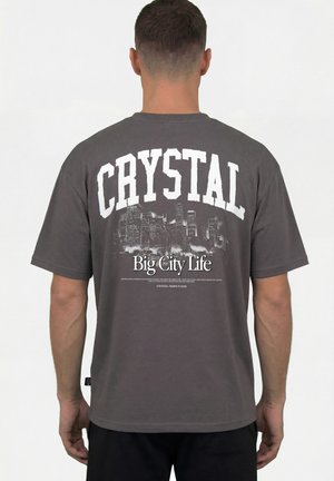 Mann trägt ein dunkelgraues T-Shirt mit einer Stadtsilhouette und dem Text "CRYSTAL Big City Life" auf dem Rücken, steht mit dem Rücken zur Kamera.