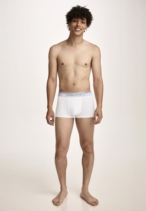 Junger Mann mit lockigem Haar, barfuß stehend, trägt weiße Jockey-Boxershorts, lächelt vor einem schlichten, hellen Hintergrund.