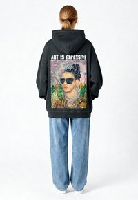 Sweat à capuche noir avec un graphisme au dos représentant une femme en lunettes de soleil accompagné du texte « L'ART EST CHER. » Porté avec un jean à jambes larges bleu clair.